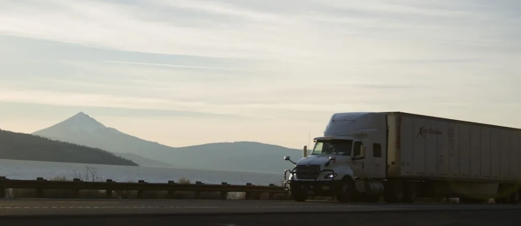 White semi-truck driving along a highway past water and mountains